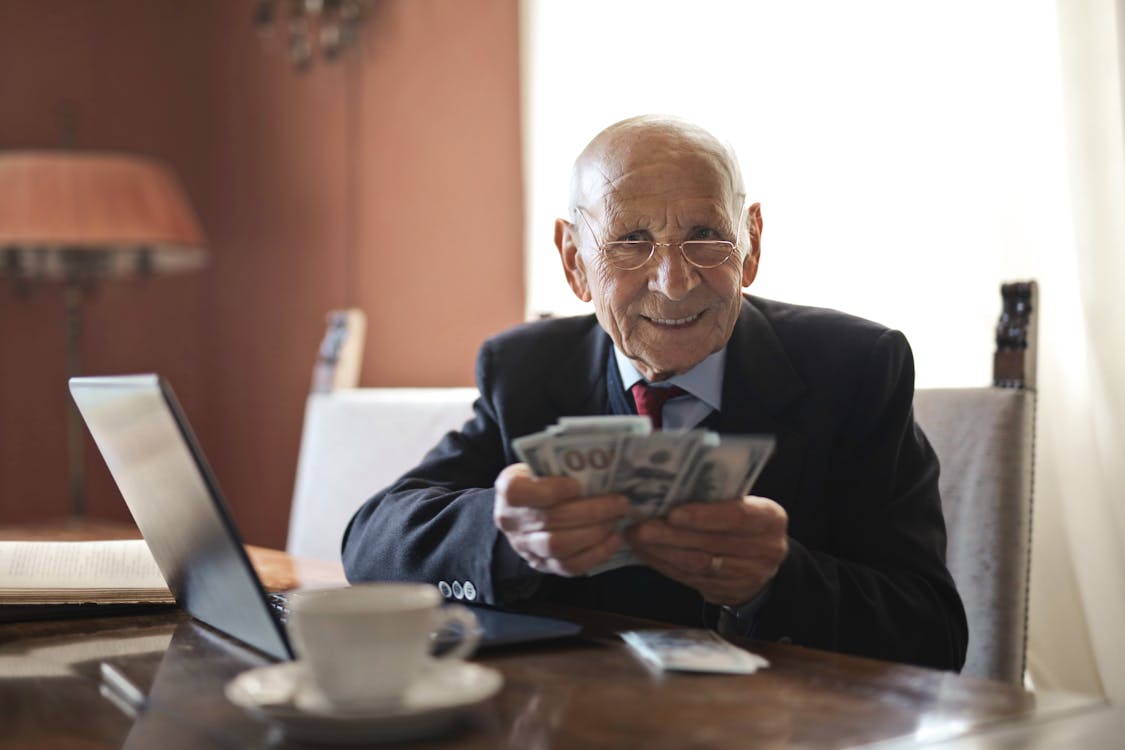 Free Confident senior businessman holding money in hands while sitting at table near laptop Stock Photo