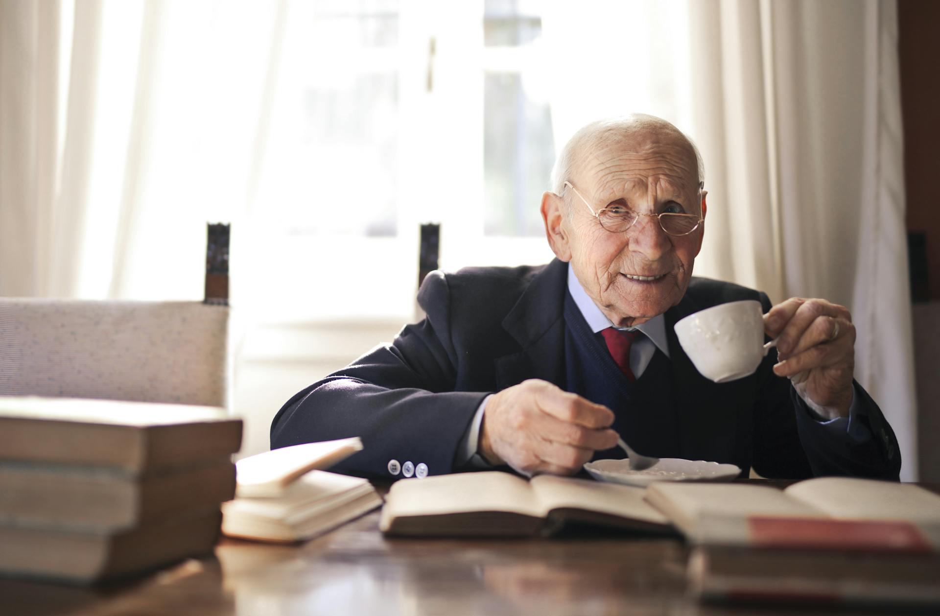 Positive senior man in formal suit and eyeglasses drinking hot beverage from white cup sitting at wooden table with books and looking at camera