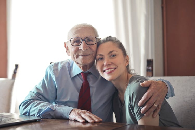 Positive Senior Man In Formal Wear And Eyeglasses Hugging With Young Lady While Sitting At Table