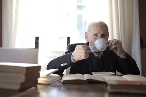Senior man drinking hot beverage while sitting at table with books