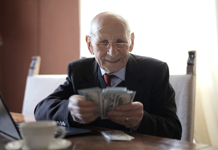 Happy Senior Businessman Counting Money While Sitting At Table With Laptop