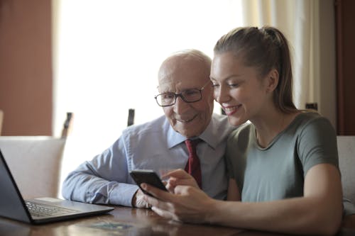 Young positive lady showing photos on smartphone to senior man while sitting at laptop