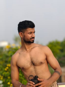 A shirtless young man poses confidently outdoors holding a dumbbell, exuding strength and vitality.