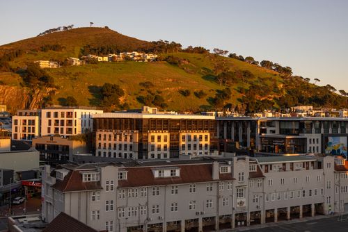 Free Golden sunlight bathes urban buildings with a verdant hill in the backdrop, exuding a serene evening ambiance. Stock Photo