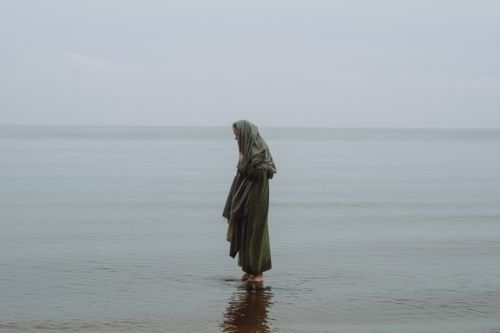 Free A woman draped in fabric stands quietly in shallow water by a calm sea, evoking serenity. Stock Photo