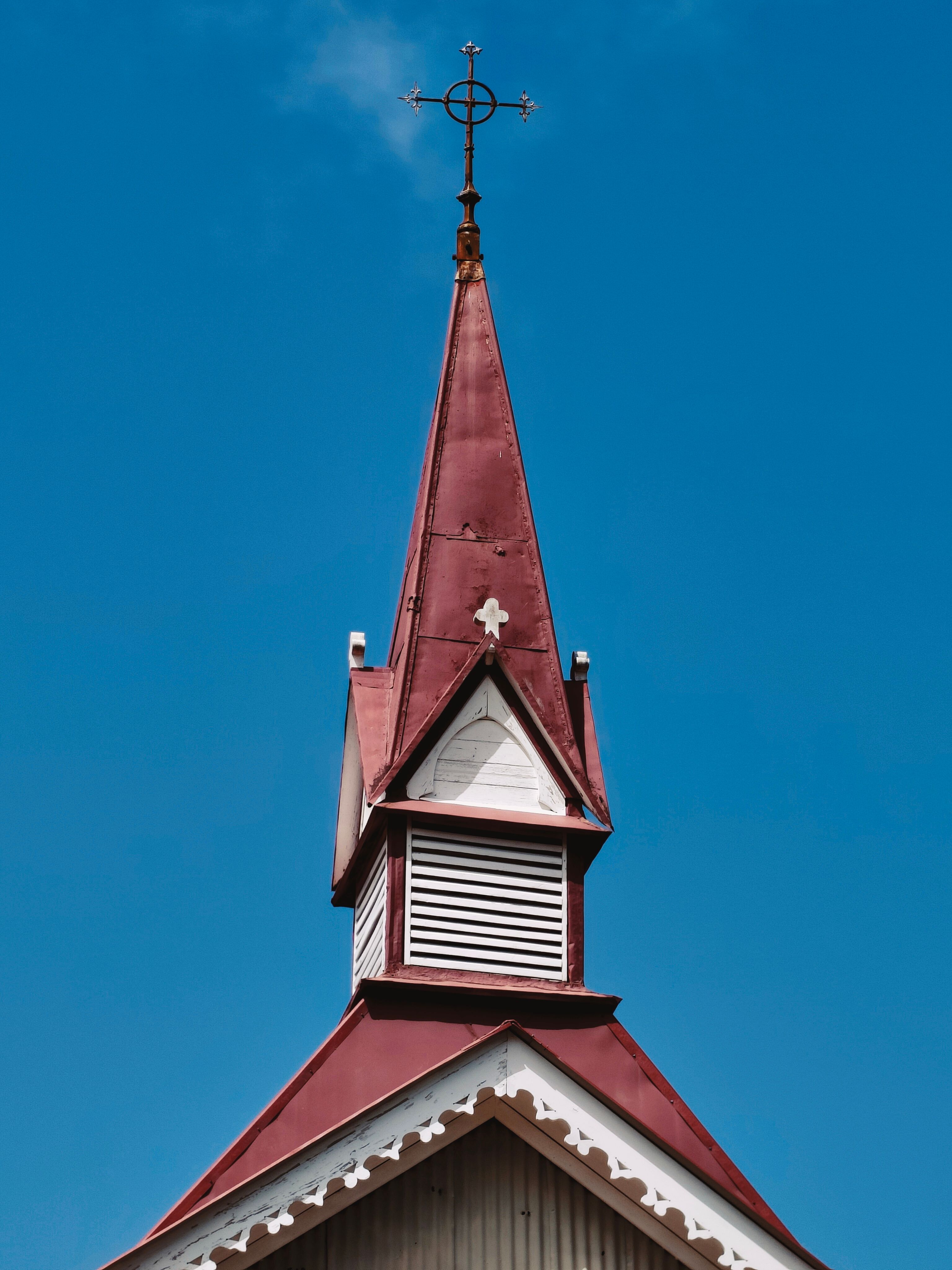 Photograph of a red church steeple with a cross against a clear blue sky, showcasing ecclesiastical architecture.