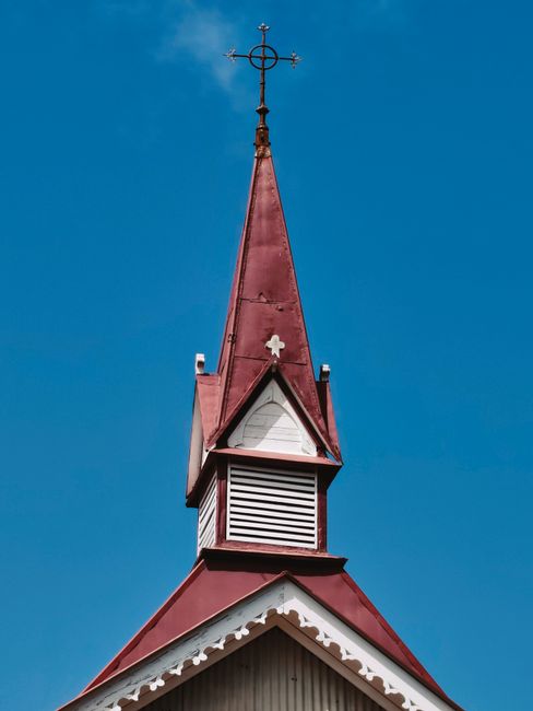 Photograph of a red church steeple with a cross against a clear blue sky, showcasing ecclesiastical architecture.