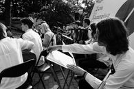 Outdoor orchestral performance with musicians playing various instruments in black and white.