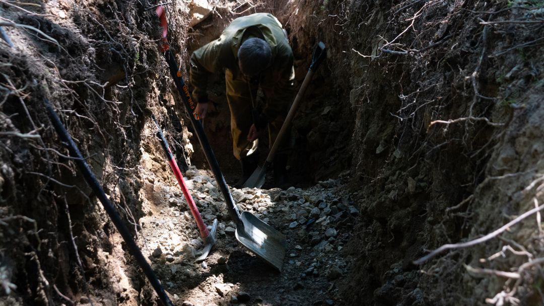 A person digging a trench outdoors with shovels and tools alongside