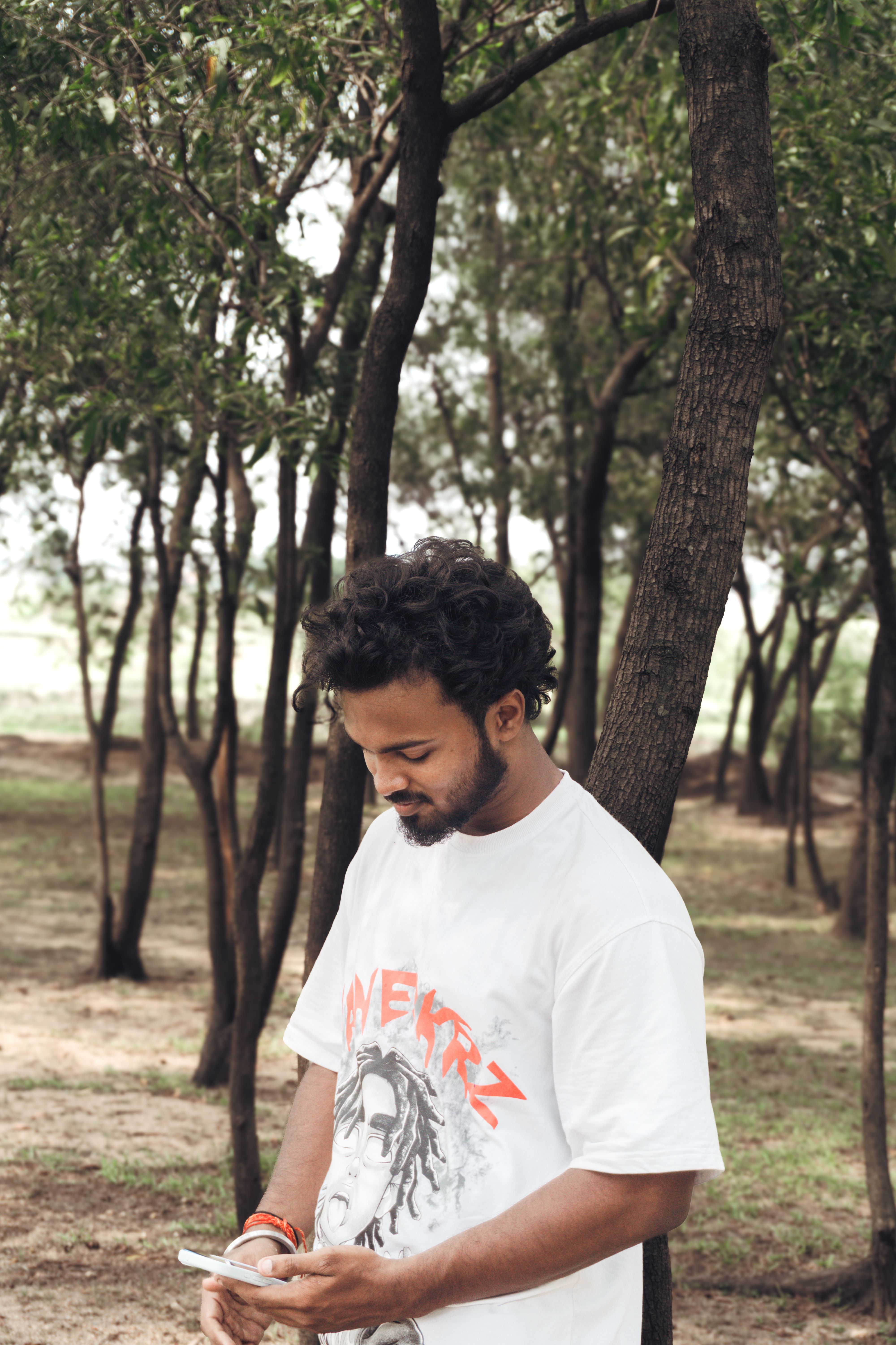 A young man standing in a forested area while using his smartphone, with trees surrounding him.