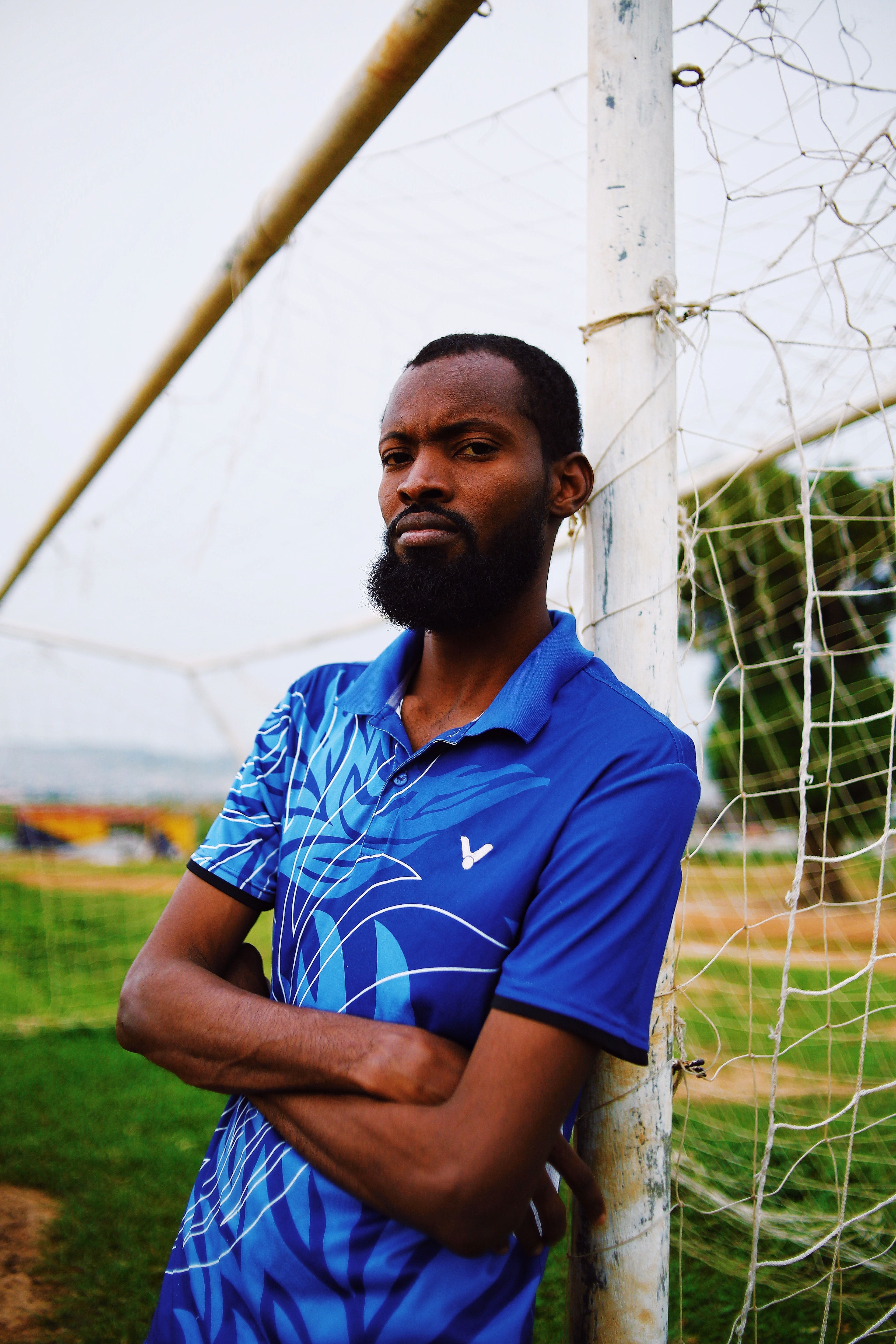 A confident Nigerian man stands by a soccer goal post in a casual blue shirt outdoors.