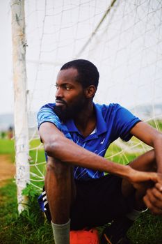 A young soccer player in blue jersey sitting by the goal post, contemplating the game.