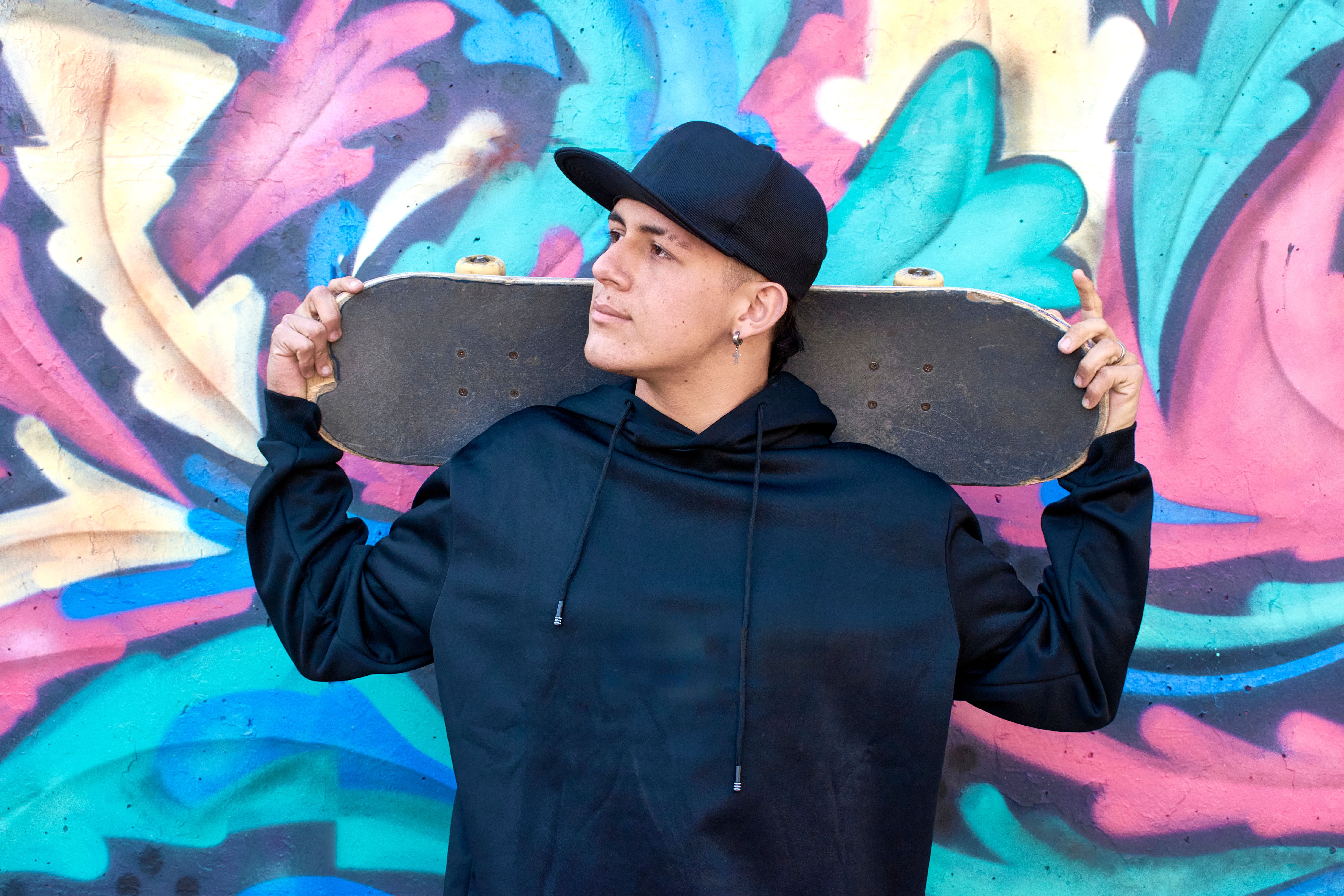 Jeune homme en hoodie noir et casquette portant un skateboard devant un mur de street art, esthetique streetwear urbaine