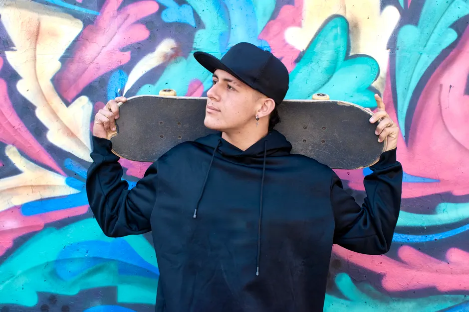 Jeune homme en hoodie noir et casquette portant un skateboard devant un mur de street art, esthétique streetwear urbaine