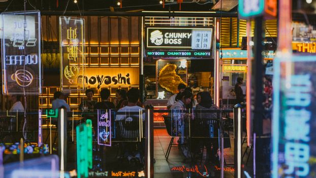 Colorful neon-lit night market scene with people dining at food stalls, creating a lively atmosphere.