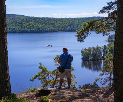 A man controls a drone over a stunning lake surrounded by lush greenery on a sunny day.