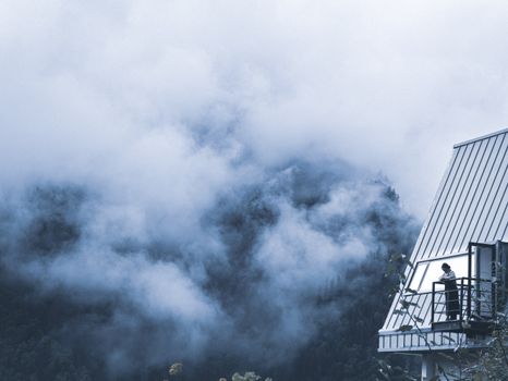 A solitary figure observes a fog-covered mountain landscape from a balcony, capturing a serene and introspective moment.