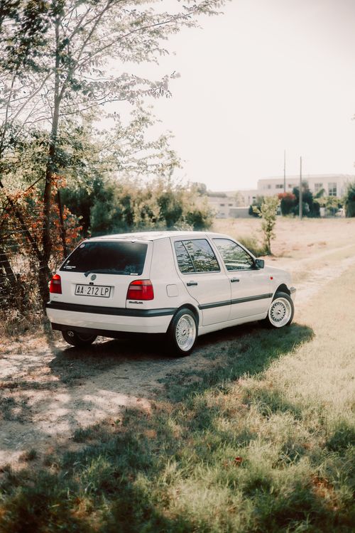 Free Vintage white hatchback car parked along a sunlit path surrounded by greenery. Stock Photo