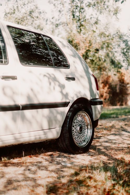 Free Vintage white car parked on a sunlit leafy path with retro wheel design, capturing nostalgic vibes. Stock Photo