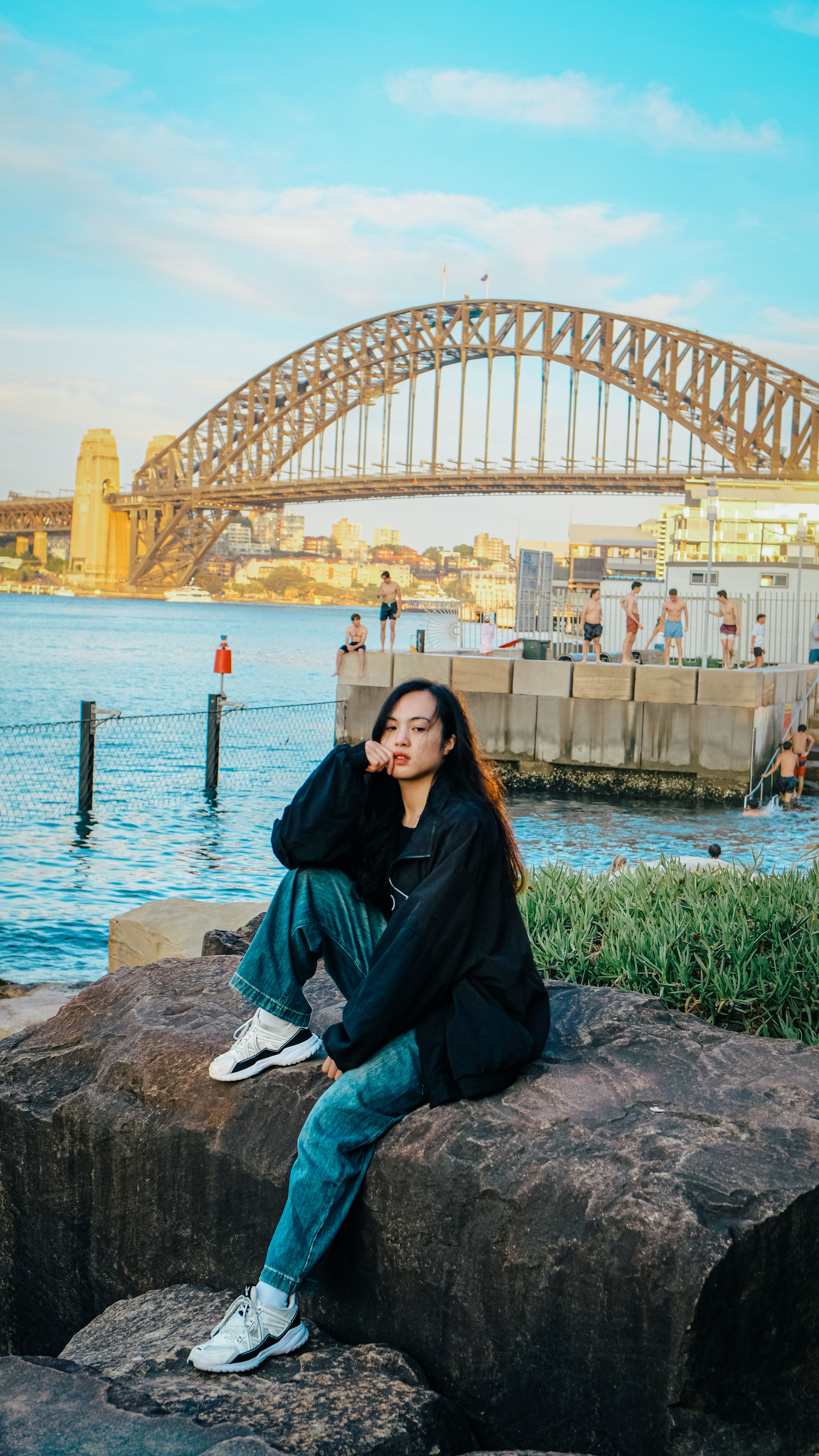 A young woman sits on rocks by the Sydney Harbour Bridge during a vibrant sunset.