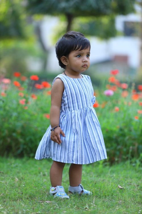 Free Young child in a striped dress standing on grass with vibrant flowers in the background. Stock Photo