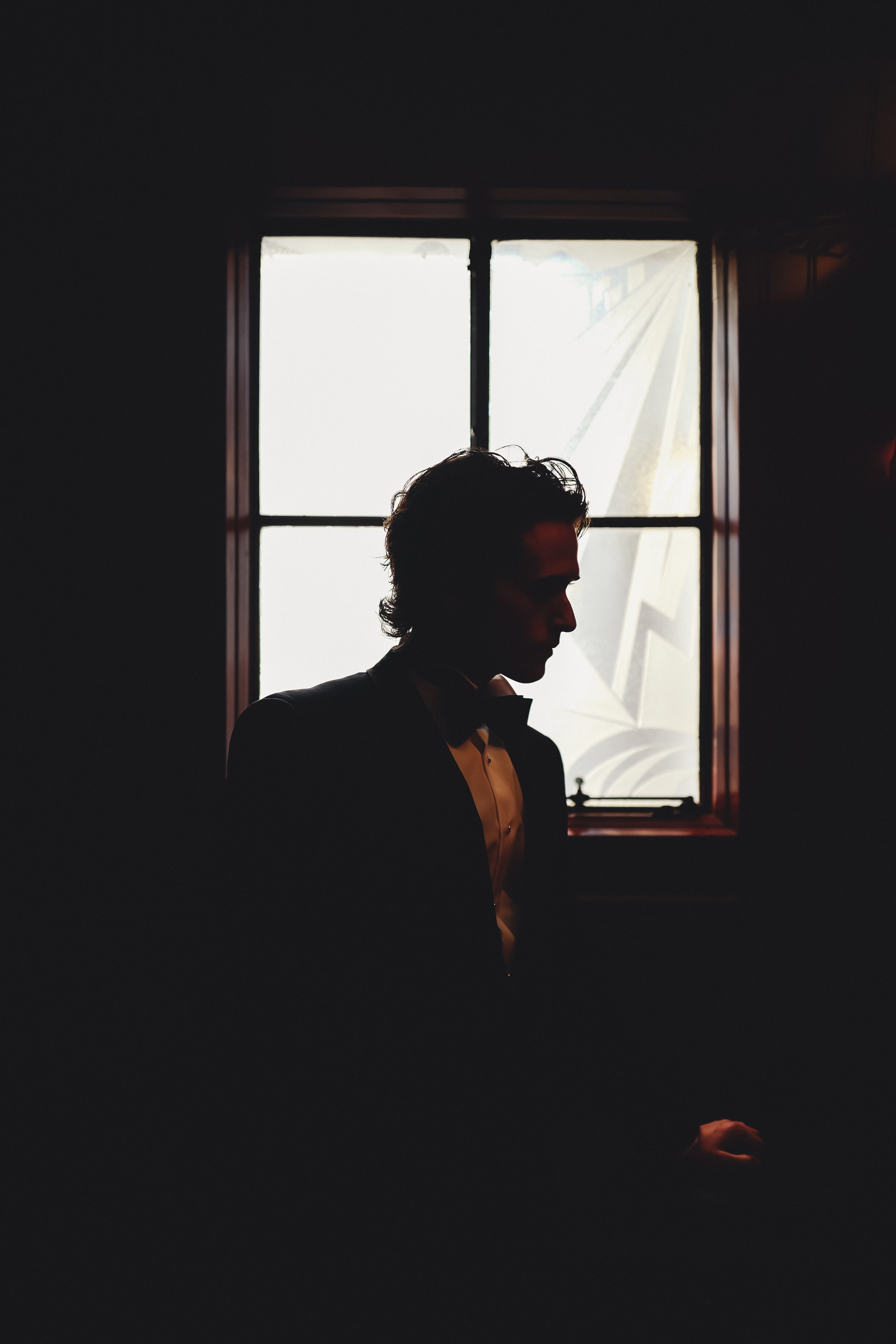 Artistic silhouette of a groom dressed in a suit, posing by a window with soft lighting, creating a dramatic scene.