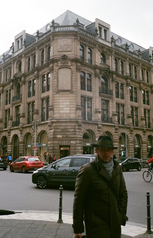 Мужчина в зеленой куртке и шляпе стоит на улице возле автомобилей и здания
