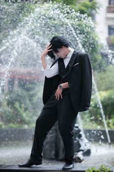A person in formal attire poses dramatically by a scenic fountain, embodying elegance.