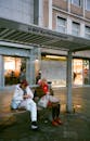 Mature women in colorful outerwear with bags sitting on bench of bus terminal with glass wall and map near modern building on pavement in town