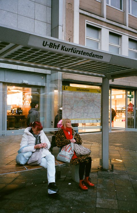 Mature women in colorful outerwear with bags sitting on bench of bus terminal with glass wall and map near modern building on pavement in town