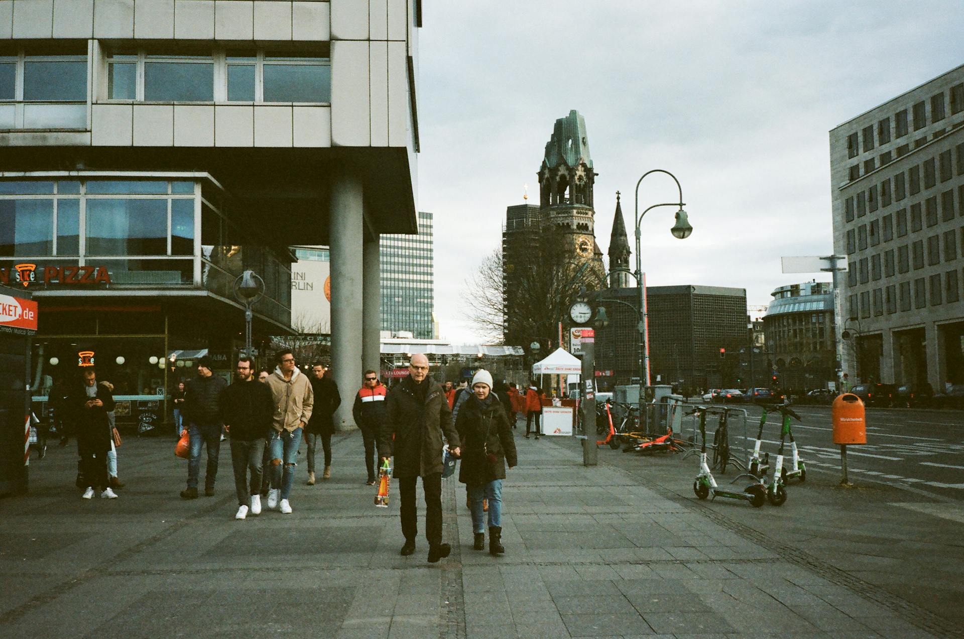 Full body of anonymous people in warm clothes walking on paved pedestrian street near modern buildings and ancient Kaiser Wilhelm Memorial Church in Berlin