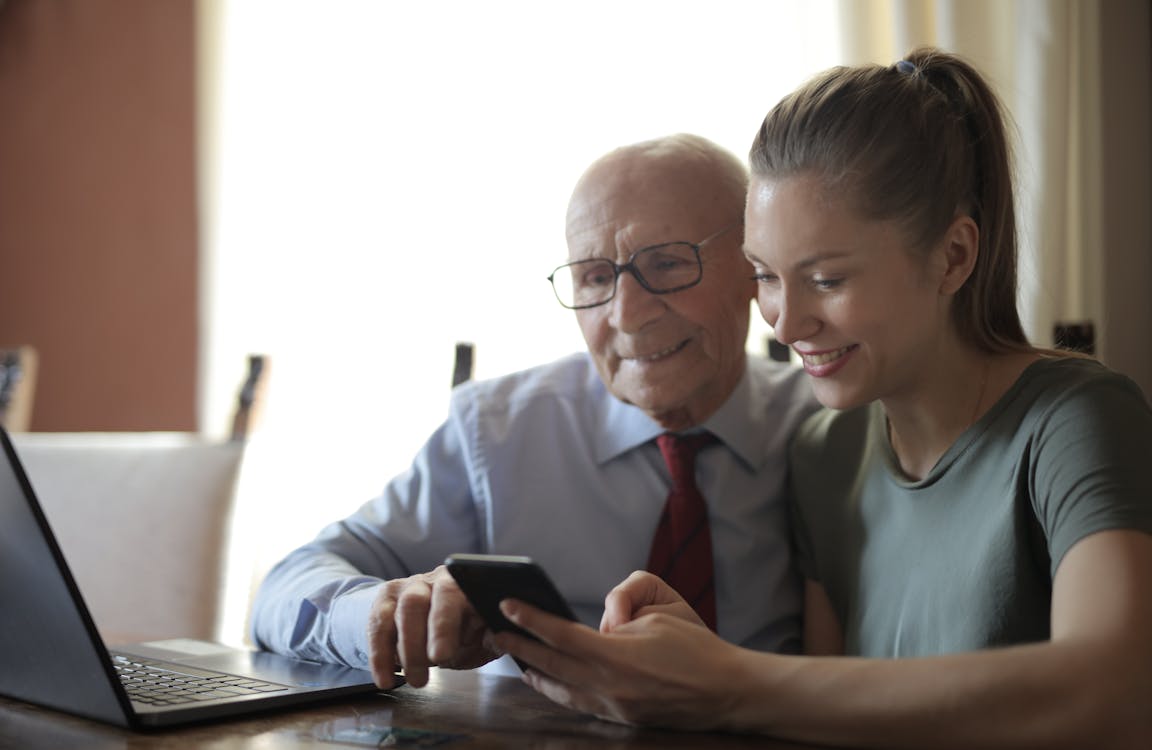 Positiver älterer Mann Und Lächelnde Junge Frau, Die Smartphone Beim Sitzen Am Tisch Beobachten