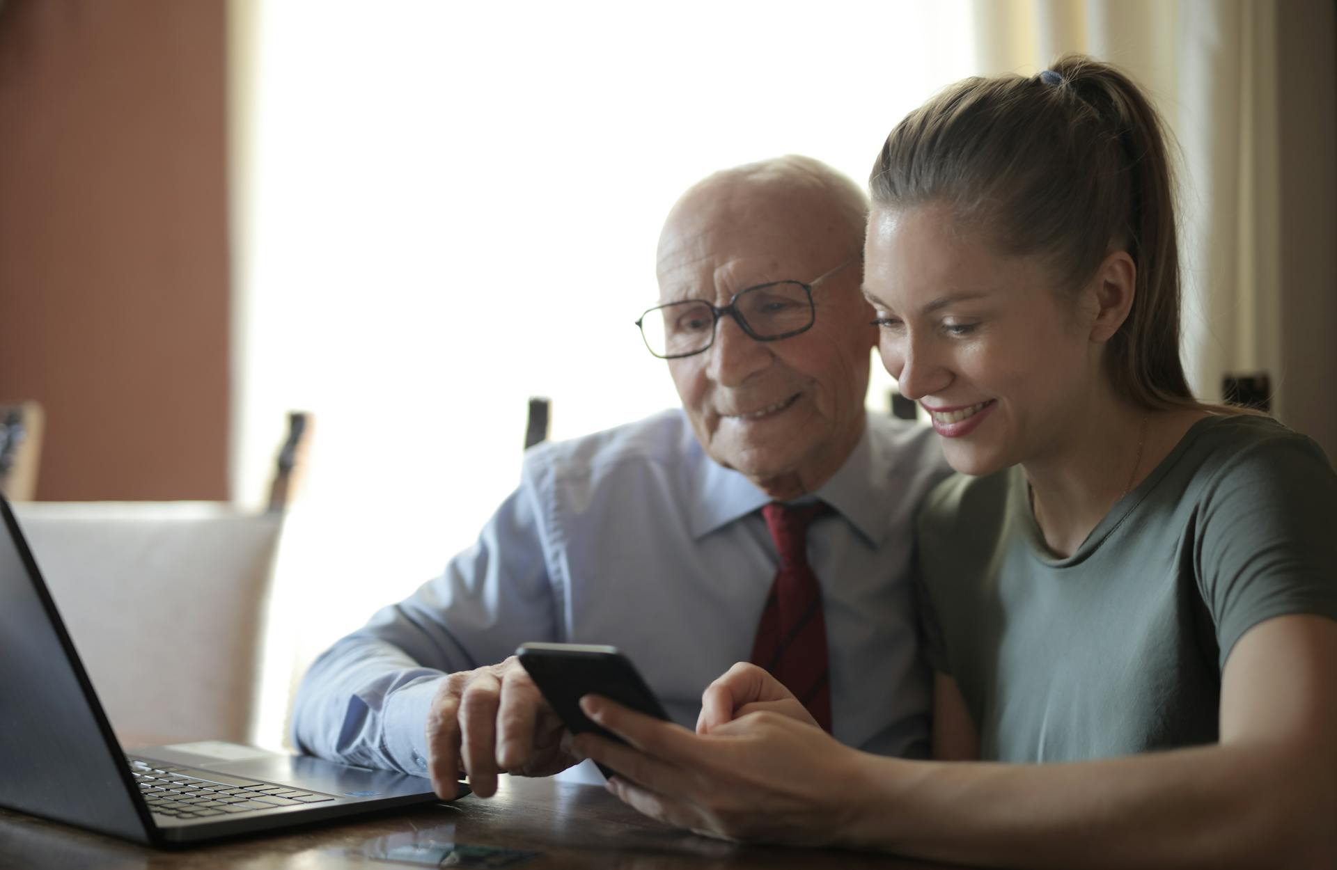 Un homme âgé positif en chemise et lunettes formelles et une petite-fille souriante partageant un téléphone portable assis près d'un ordinateur portable à table