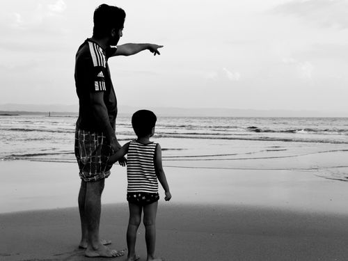 Free A father and child stand on a serene beach, gazing at the ocean in a black and white photograph. Stock Photo