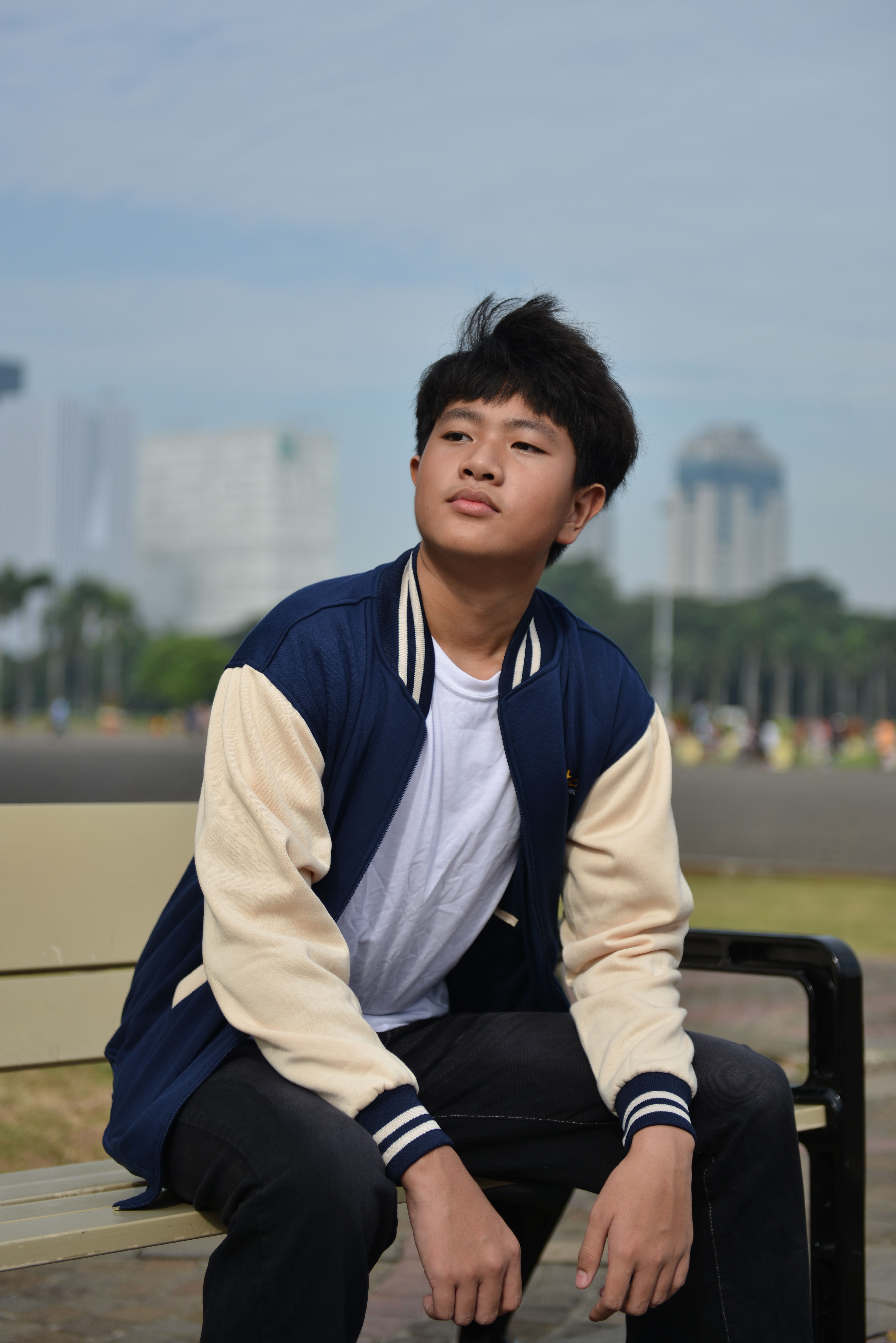 Teenager sitting on a bench outdoors, wearing casual clothing, in a city park setting.