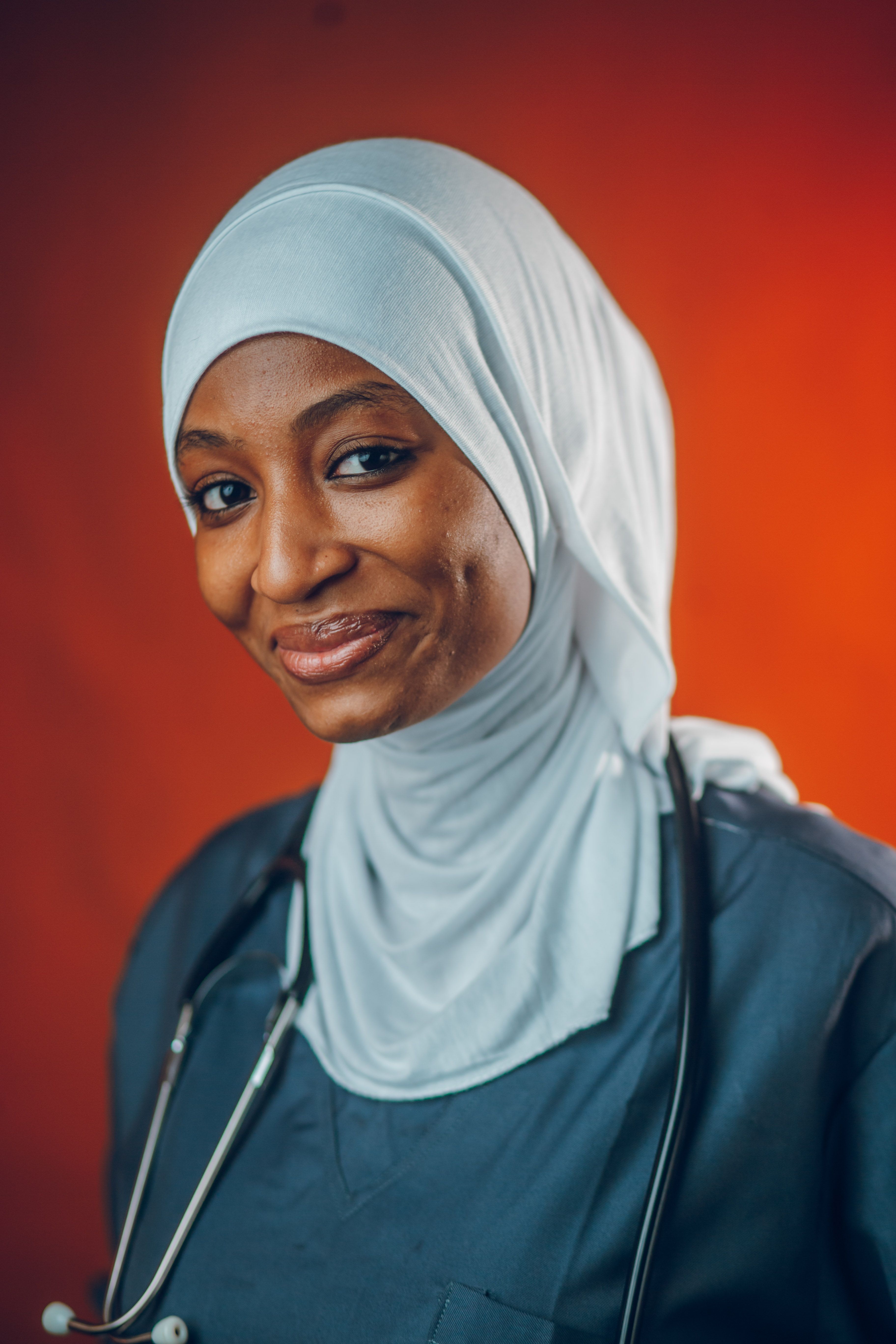 Smiling female doctor wearing a hijab and stethoscope on a vibrant orange background.