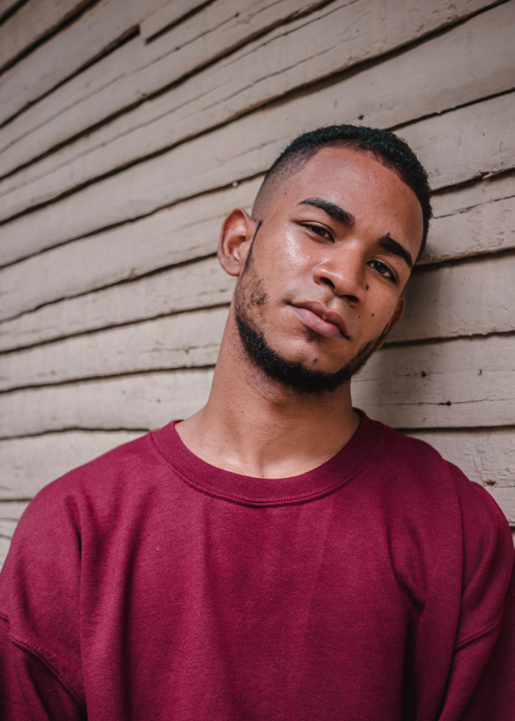 Serious young ethnic bearded male in casual clothes leaning on wooden wall and looking at camera pensively