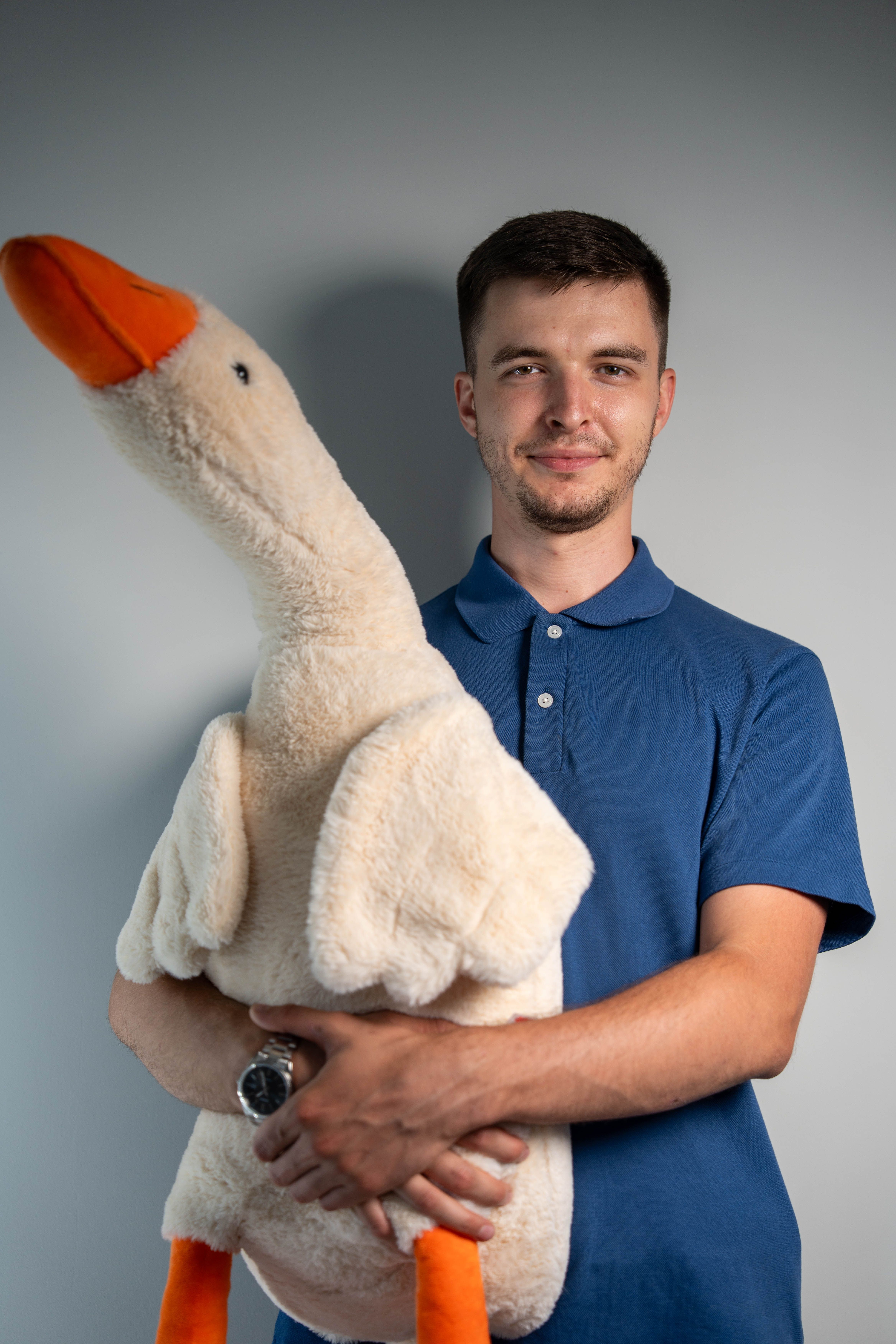A young man in a blue shirt holding a large plush goose indoors with a neutral background.