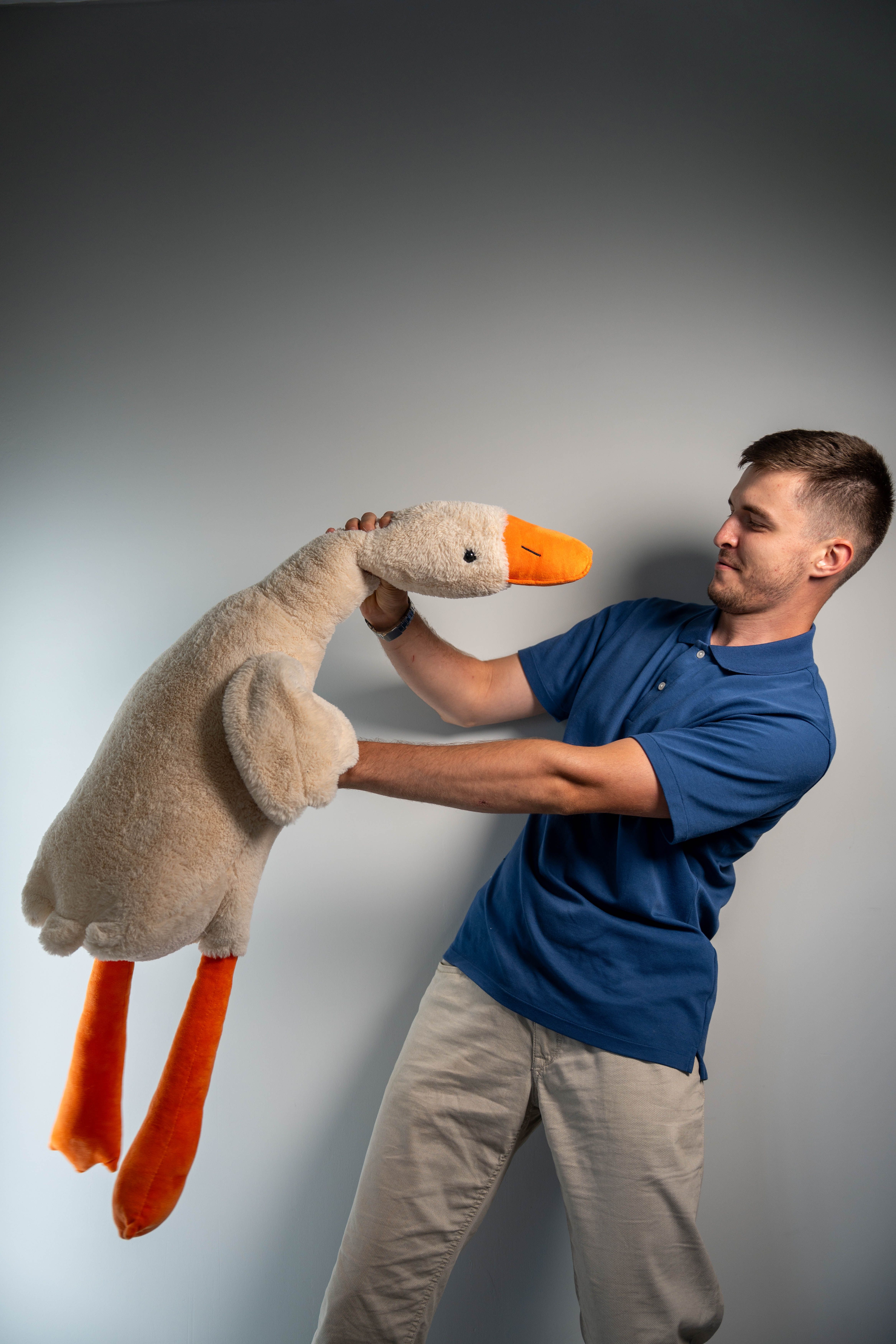 Young man in blue shirt lifting a giant stuffed duck toy against a plain backdrop.