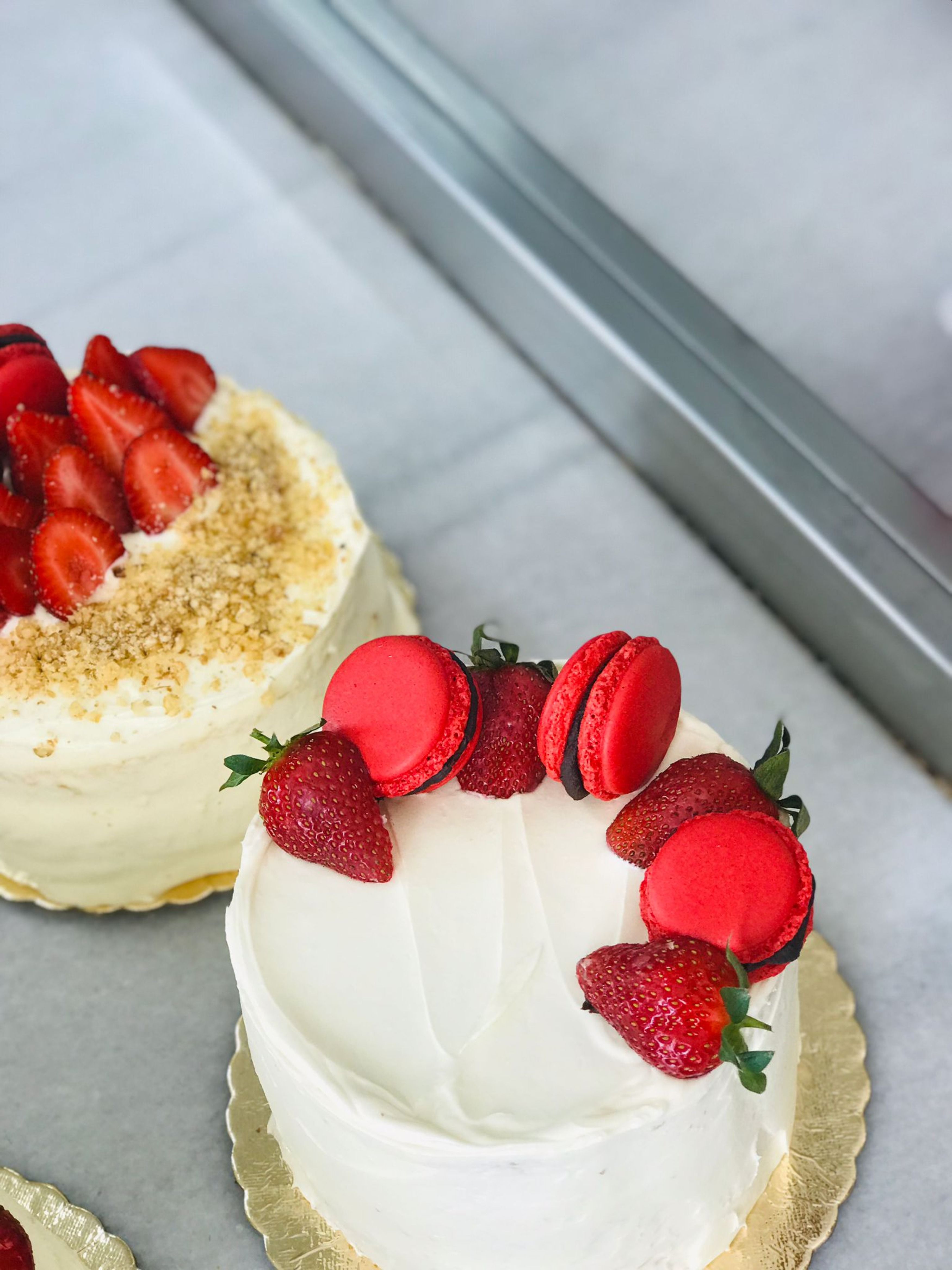 Two cakes topped with strawberries and macarons on a display counter.
