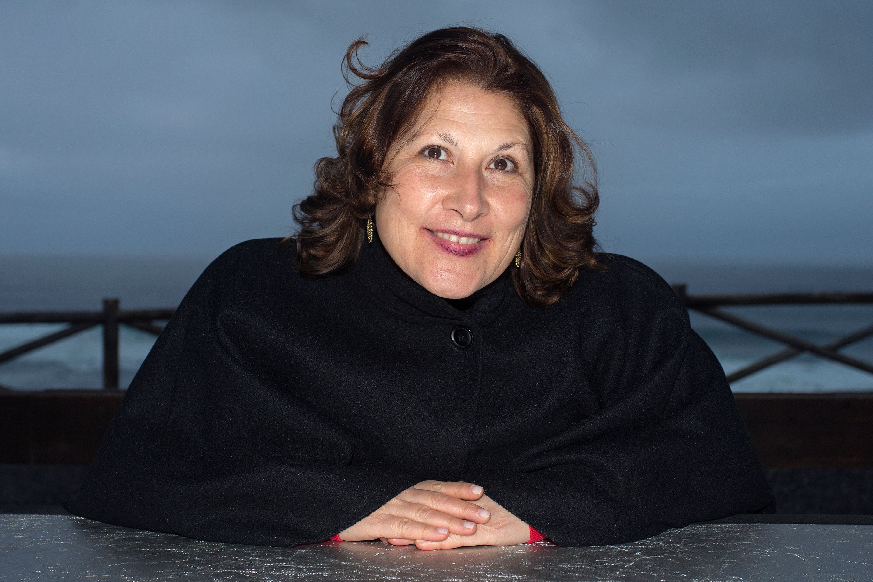 Woman in a black coat smiles warmly against a seaside backdrop.