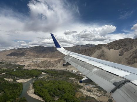 A stunning aerial shot of Leh, featuring an airplane wing over lush valleys and rocky mountains under a blue sky.