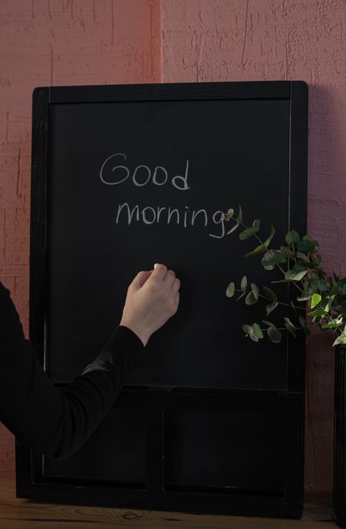 Free A hand writes 'Good morning' on a chalkboard with a cozy, plant-filled backdrop. Stock Photo
