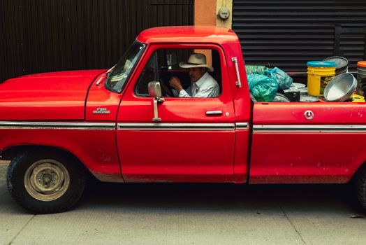 A classic red truck with a driver wearing a hat, parked in an urban area with colorful containers.