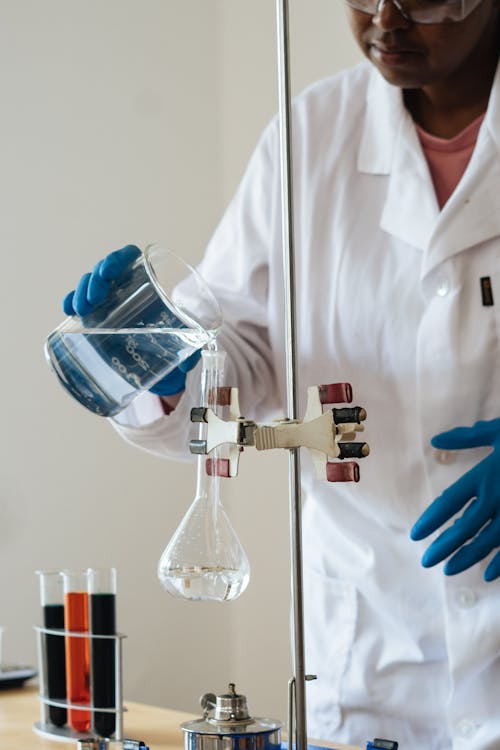 Crop African American laboratory technician in protective goggles pouring transparent fluid into flask while standing in front of colorful test tubes with reagents