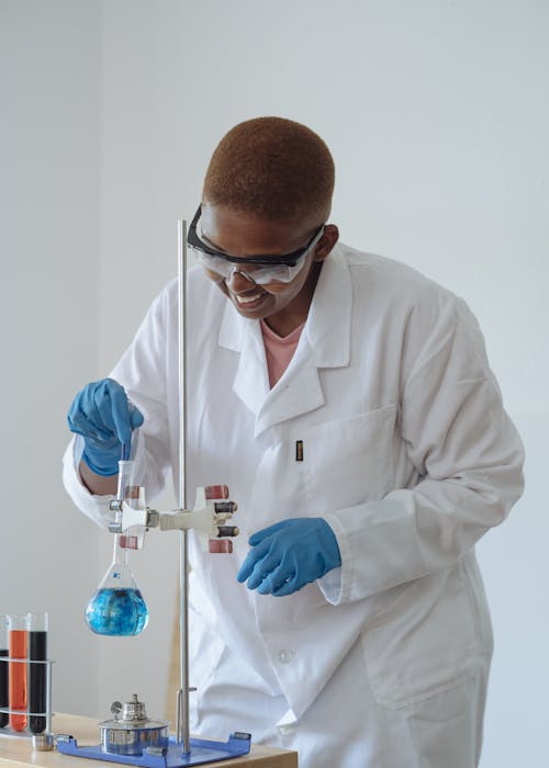 Positive African American student in protective goggles stirring light blue liquid in glassware with medical pipette while conducting experiment in science center