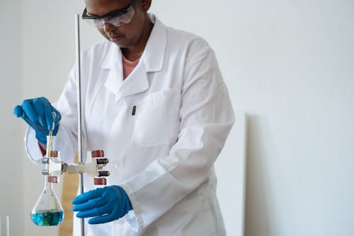 Serious laboratory technician dressed in uniform and safety glasses carrying out titration during work in laboratory inserting pipette into flask with reagent
