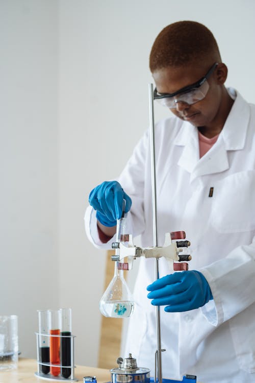 Serious scientist in uniform investigating sample while mixing reagents from different test tubes in flask mounted on stand in research center