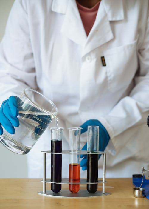 Crop of faceless researcher in uniform checking chemical reaction by pouring transparent liquid to test tubes with colored mixtures