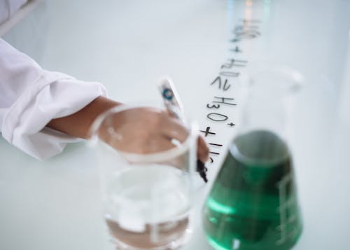 Body part of unrecognizable scientist in white uniform writing down formula after providing chemical research with fluid in flask during science lesson in university